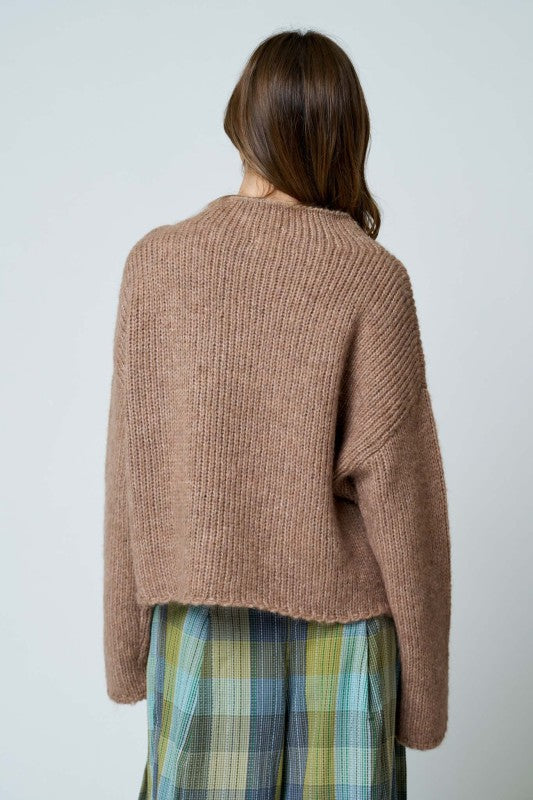 Person wearing a brown knitted sweater and plaid pants against a light background