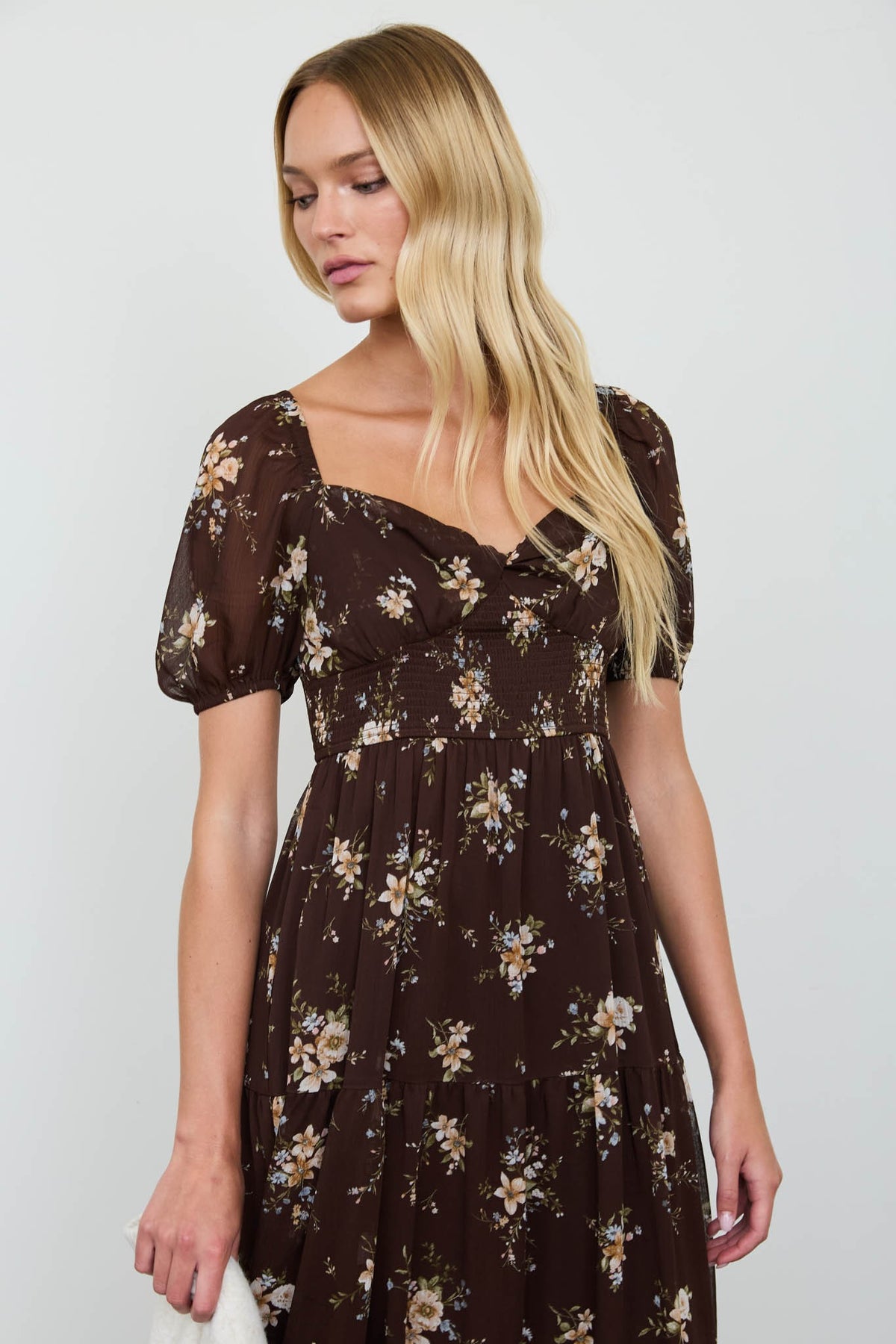 A blonde woman wearing a dark brown tiered midi dress with a white and yellow floral print, puff sleeves, and a square neckline, photographed against a light gray studio background.