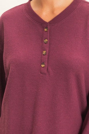 A close-up of a ribbed knit henley top in a deep berry or plum purple color, featuring a three-button placket with gold-toned buttons.