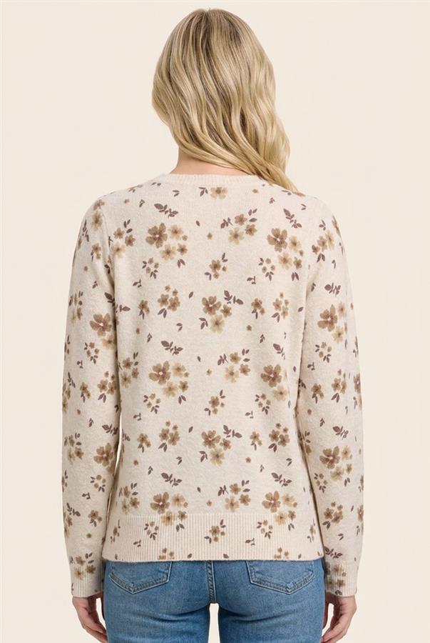 A woman viewed from the back wearing a cream-colored knit sweater with a brown and beige floral pattern, paired with blue jeans, against a solid light beige background.