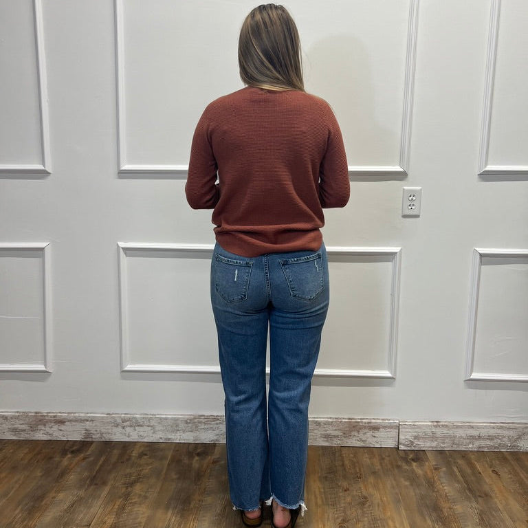 A woman viewed from behind wearing a rust-brown long-sleeve knit sweater and blue jeans, standing on a wood floor against a white paneled wall.