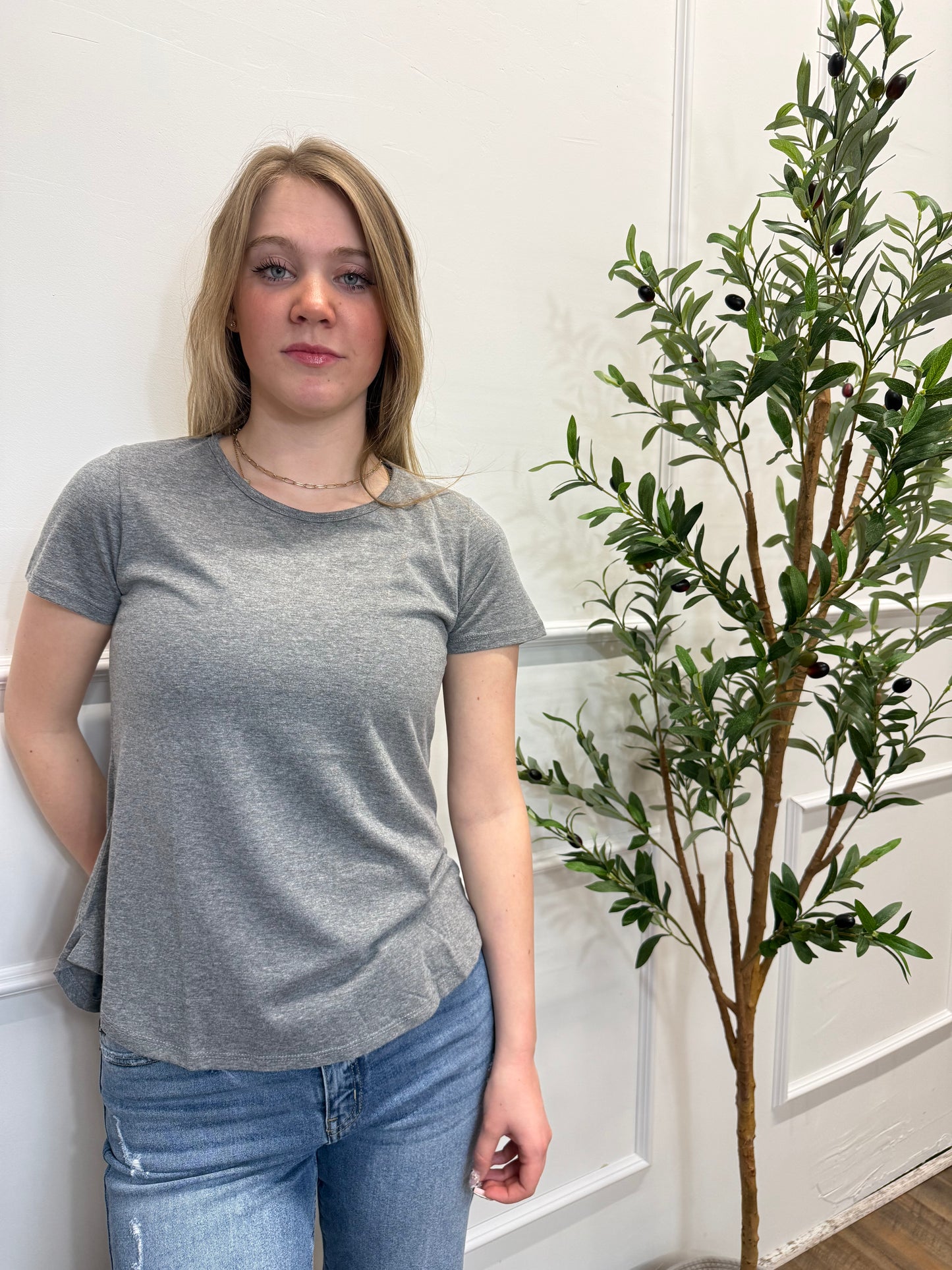 Person wearing a gray shirt and blue jeans standing next to a potted plant indoors.