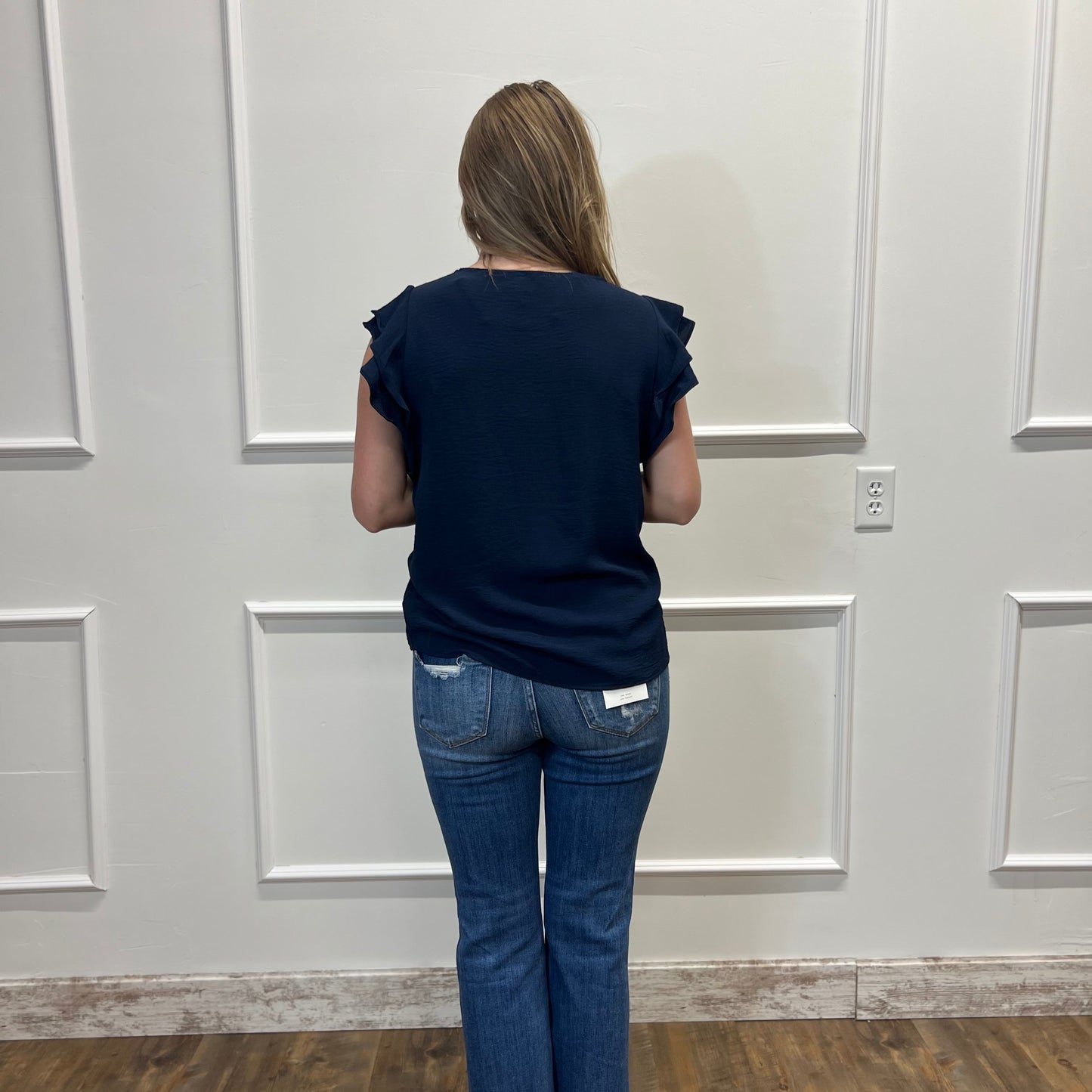 Person wearing a navy blue top and jeans standing against a white paneled wall.