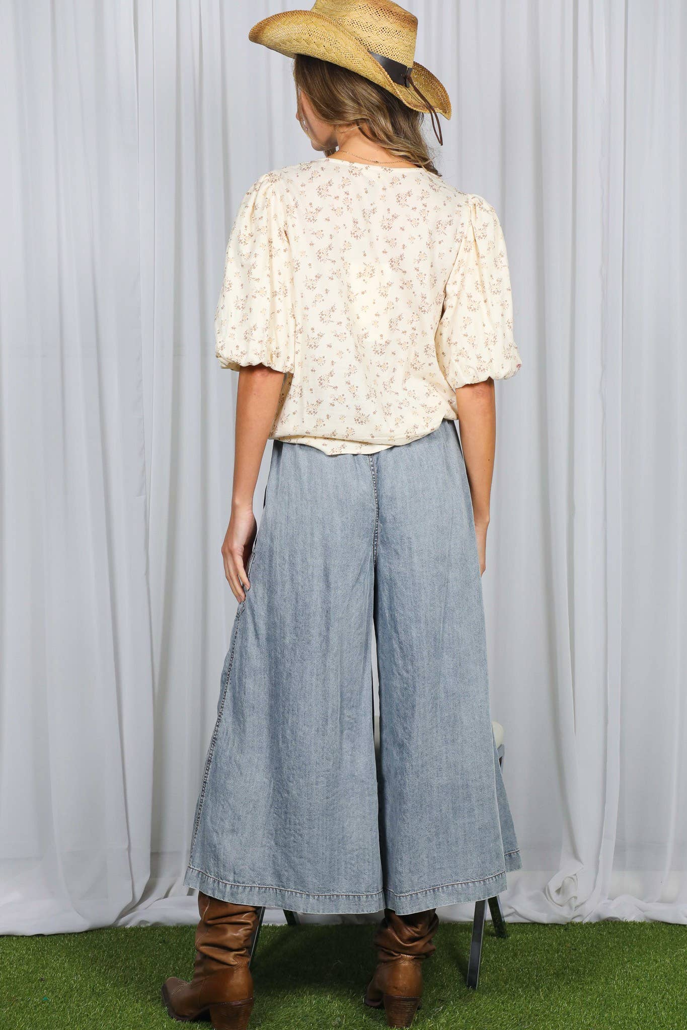 Person wearing a floral blouse, wide-leg jeans, and brown boots with a straw hat against a white curtain background.