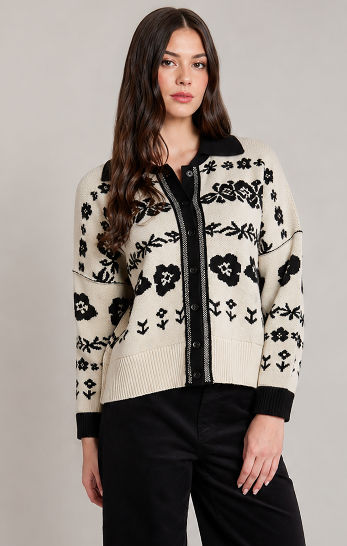 A woman modeling a cream-colored knit cardigan with black floral patterns and black trim, paired with black pants, against a solid light gray studio background.