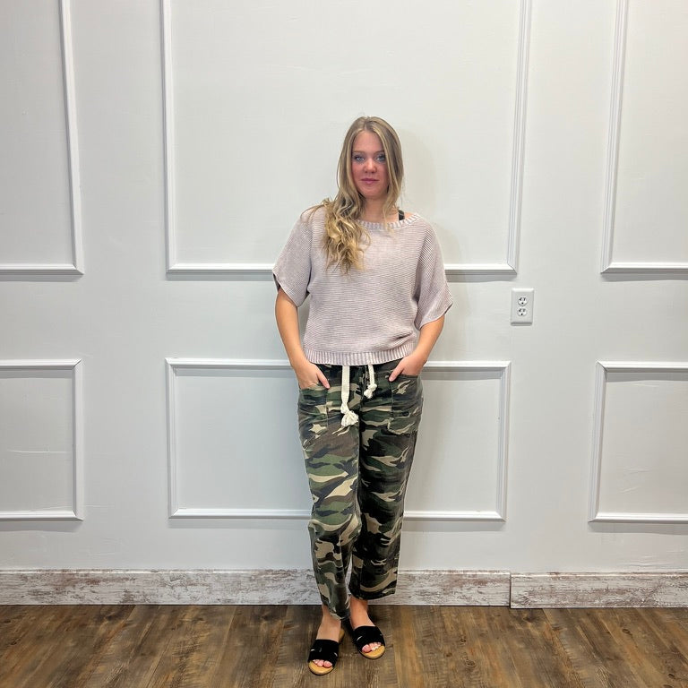 A woman stands against a white paneled wall on a wood floor, wearing a light beige short-sleeve top and camouflage drawstring pants with black slide sandals.