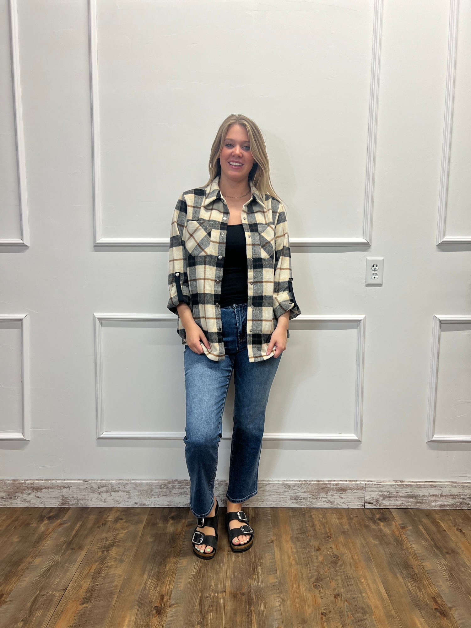 A woman stands against a white paneled wall wearing a black, cream, and gray plaid button-up shirt over a black top, paired with blue jeans and black sandals.