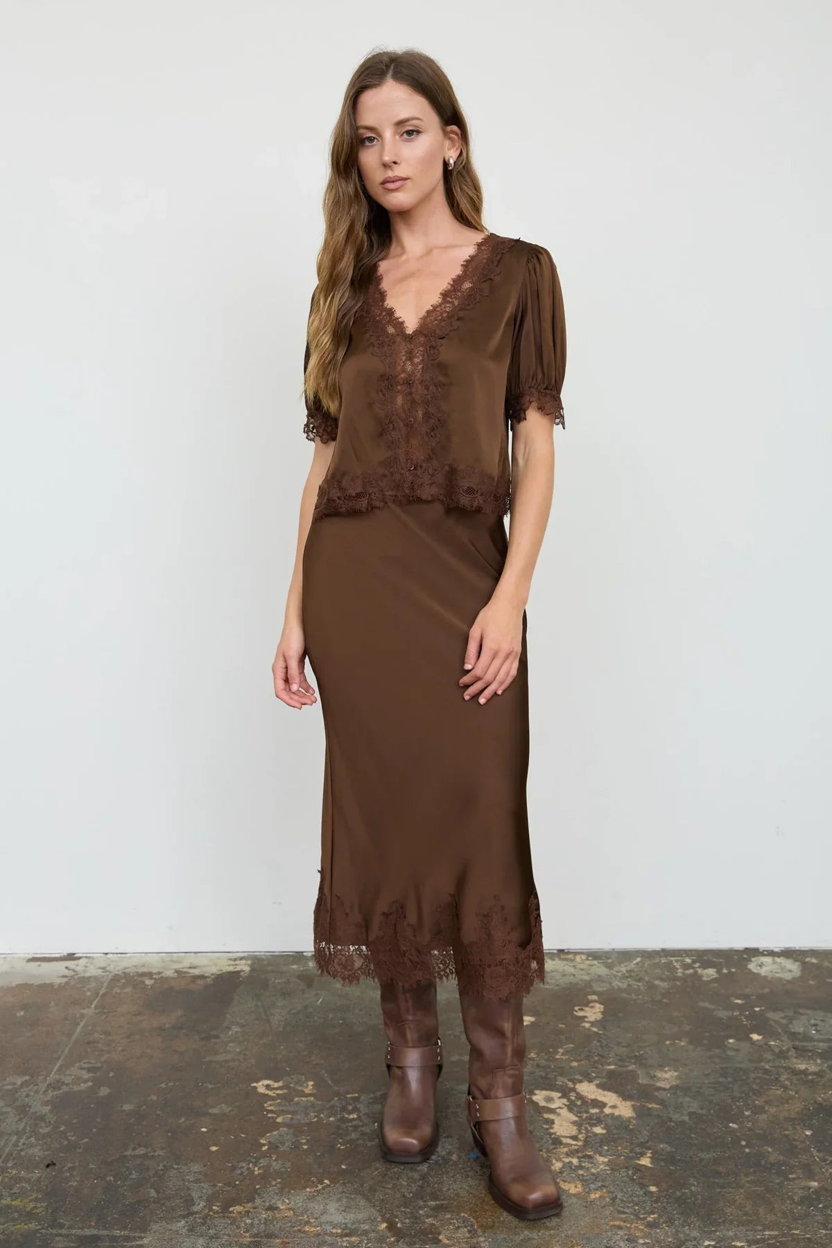 A woman modeling a brown satin midi dress with lace trim and short puff sleeves, paired with brown leather boots, against a white studio wall.