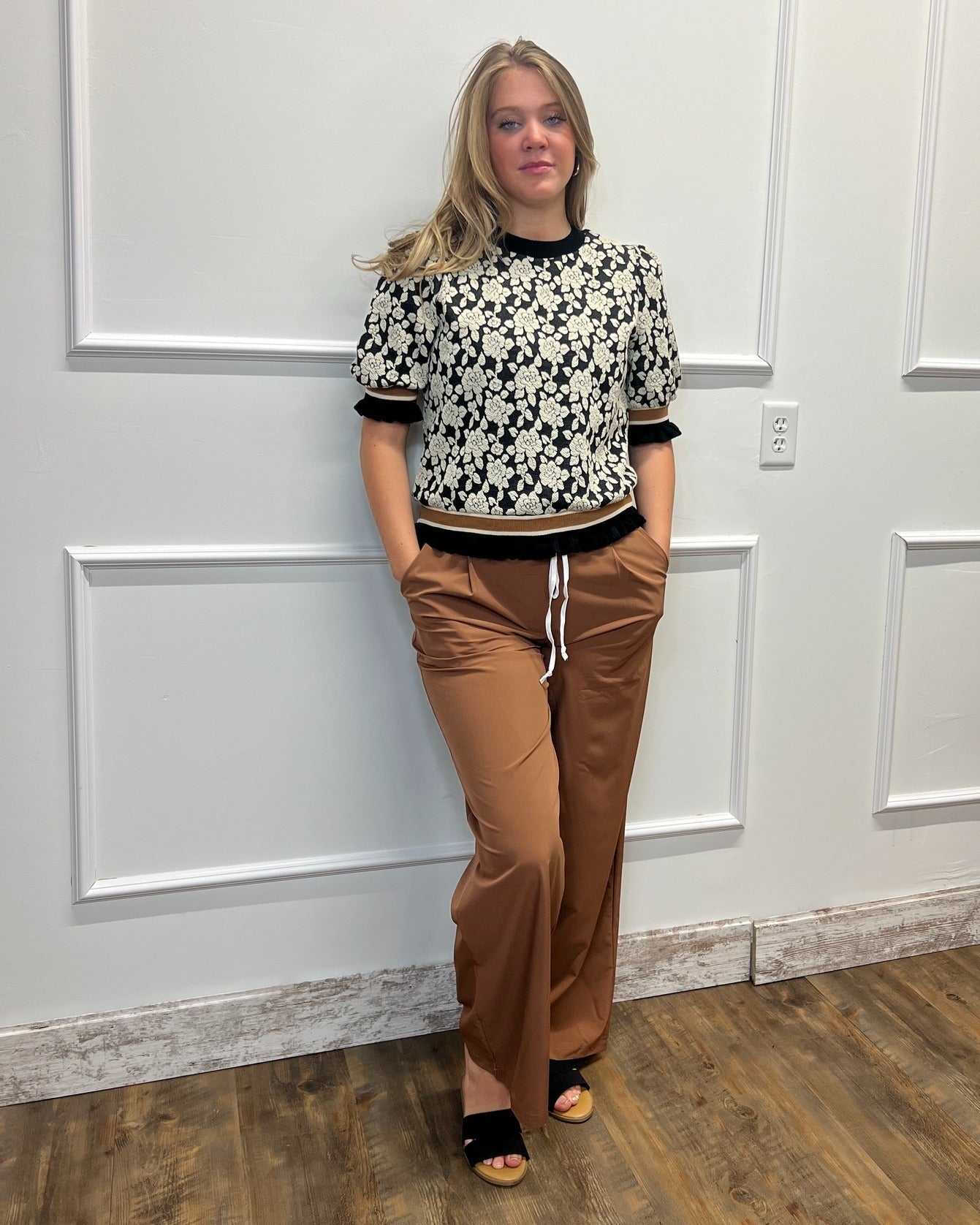 A woman stands indoors against a white paneled wall wearing a black-and-white floral short-sleeve top with black trim, brown wide-leg pants with a white drawstring, and black sandals.