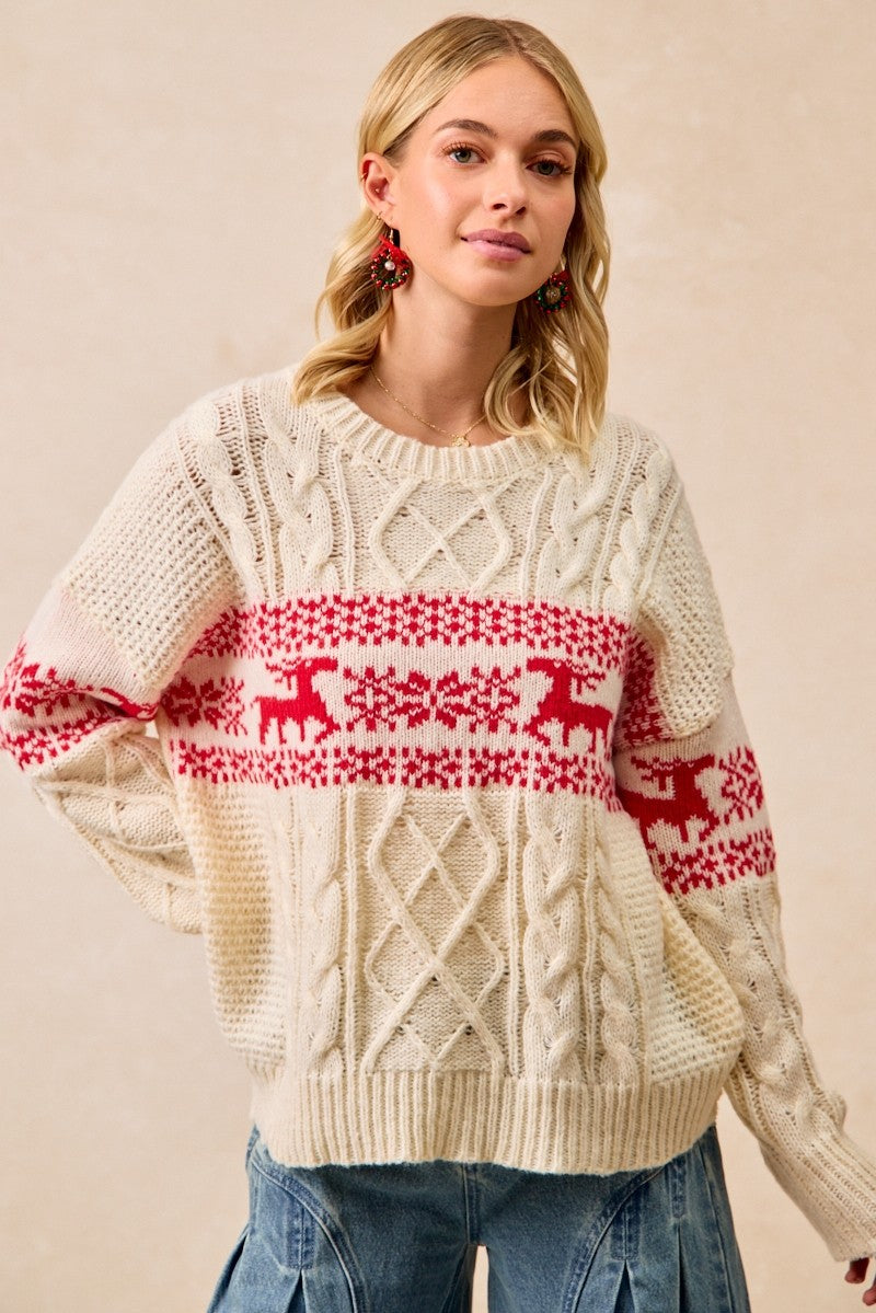 A woman wearing a cream-colored cable-knit sweater with a red horizontal band featuring reindeer and snowflake patterns, paired with blue jeans, set against a plain beige background.
