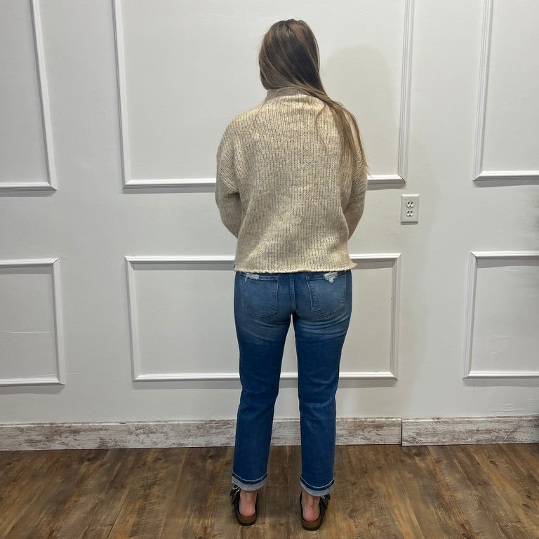 A woman viewed from behind wearing a textured cream knit sweater, blue jeans, and brown heels, standing in front of a white paneled wall on a wood floor.