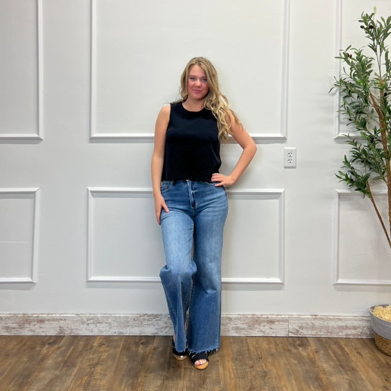 Woman in black tank top and blue jeans standing against a white paneled wall.