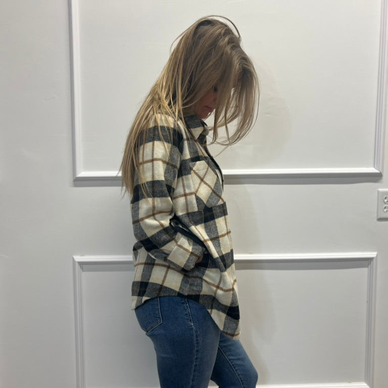 A woman with blonde hair is shown from the side, wearing a black and cream plaid flannel shirt and blue jeans, standing in front of a white paneled wall.