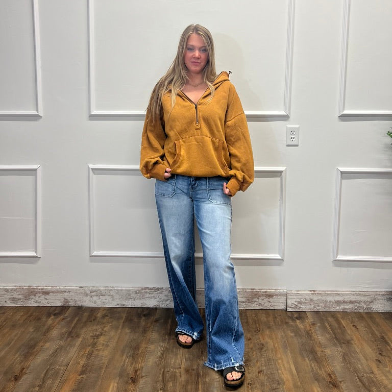 A woman stands in a room with white paneled walls and a wood floor, wearing a mustard yellow zip-up hoodie and light blue wide-leg jeans.