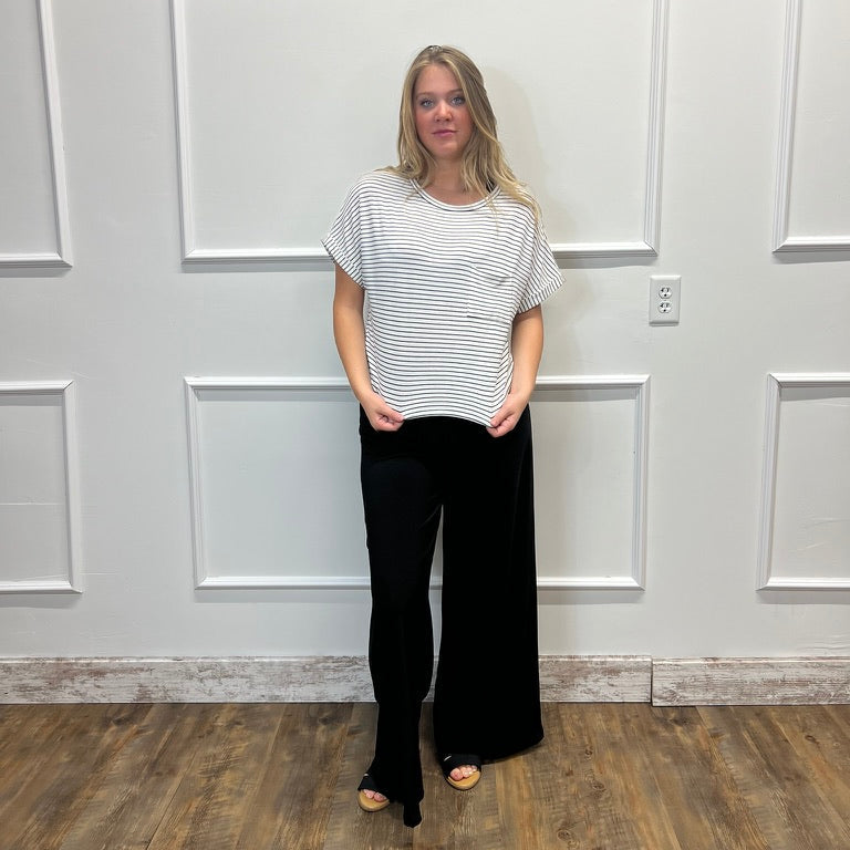 A woman stands in a room with white paneled walls and a wood floor, wearing a white short-sleeve top with thin black horizontal stripes and black wide-leg pants.