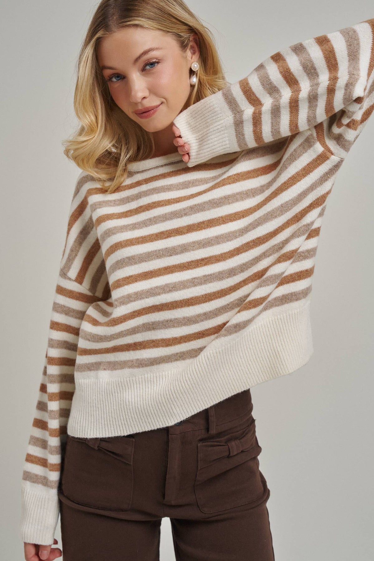 A woman wearing a cream and brown horizontally striped knit sweater with a wide crew neck and ribbed cuffs, paired with dark brown pants, against a solid light gray background.