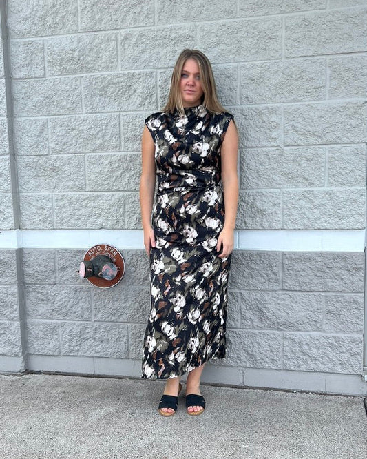 A woman stands outdoors against a light gray brick wall wearing a sleeveless black midi dress with a white floral pattern and dark sandals.