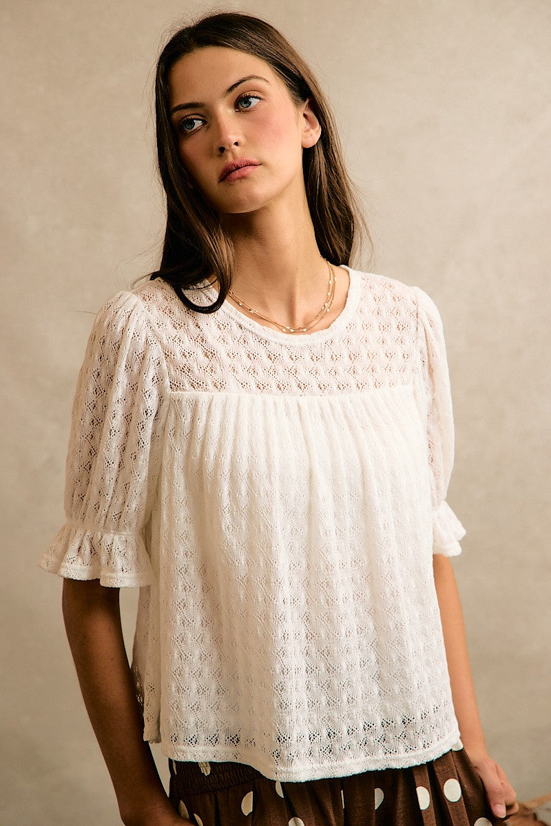 A woman wearing a white eyelet lace short-sleeve blouse with a gathered bust and ruffled sleeves, paired with a brown and white polka dot skirt, set against a neutral textured beige background.
