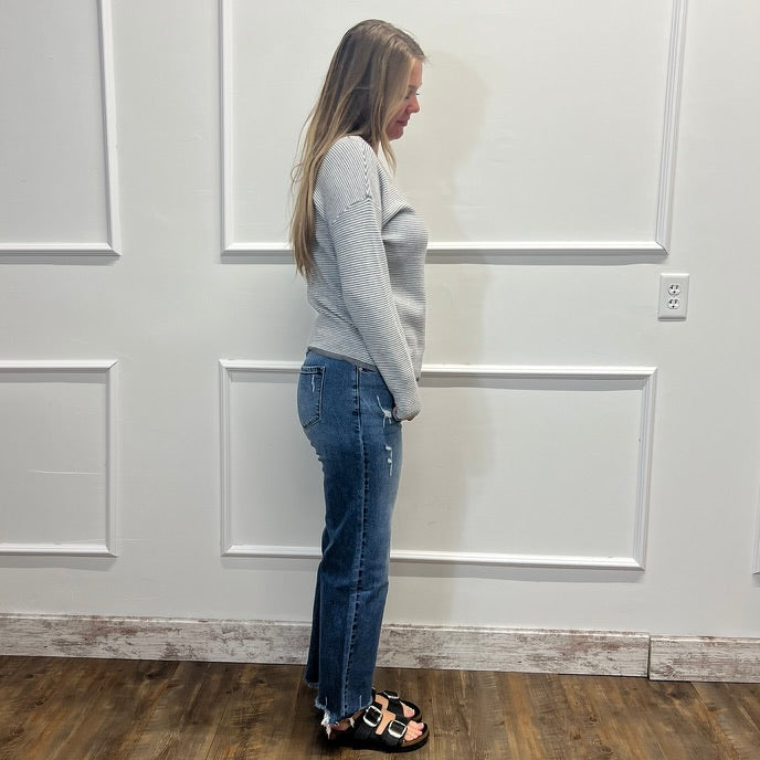 A woman stands in profile wearing a light gray long-sleeve knit top, blue jeans, and black slide sandals with a leopard print strap, set against a white paneled wall and wood-look floor.