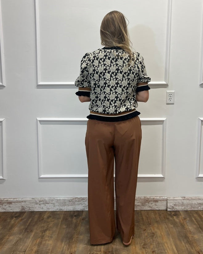 A woman viewed from behind wearing a black and white floral patterned short-sleeve top with black trim and brown wide-leg pants, standing in front of a white paneled wall.