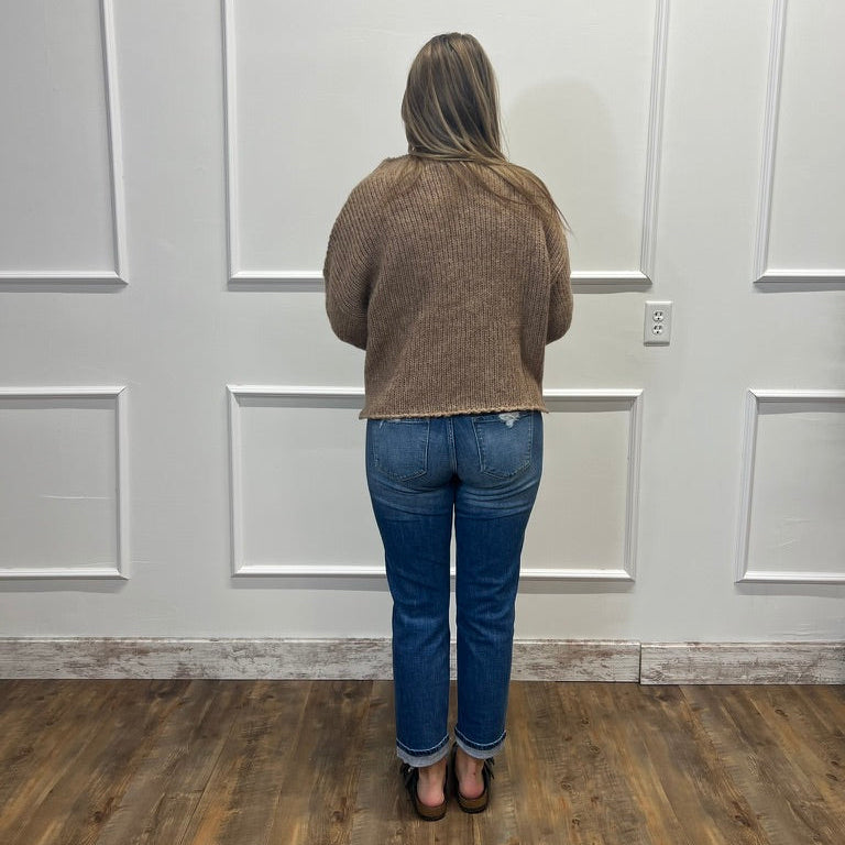 A woman viewed from behind wearing a brown ribbed knit sweater and blue jeans, standing on a wood floor in front of a white paneled wall.