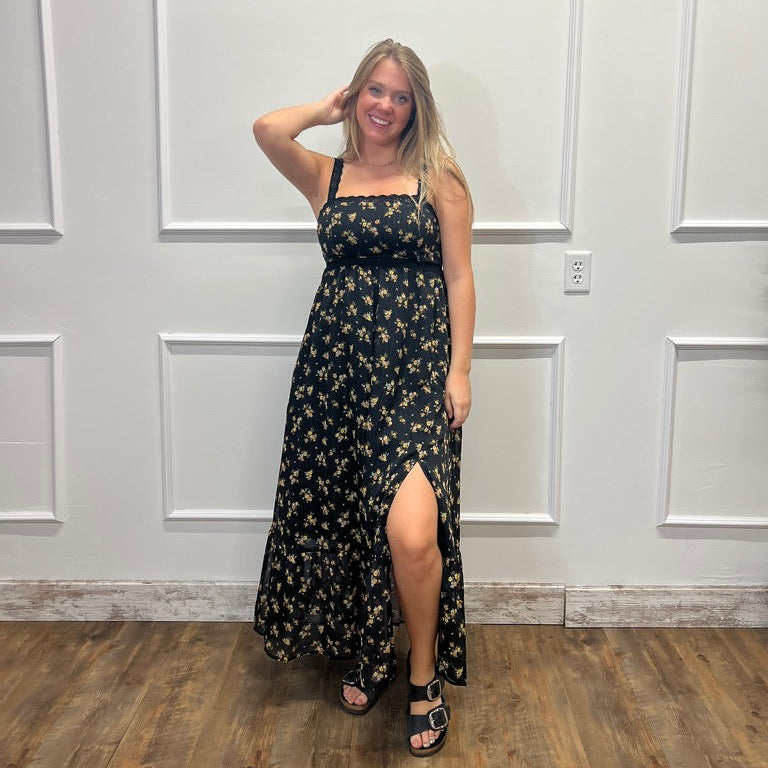 A woman modeling a black midi dress with a yellow floral pattern, standing in front of a white paneled wall on a wood floor.