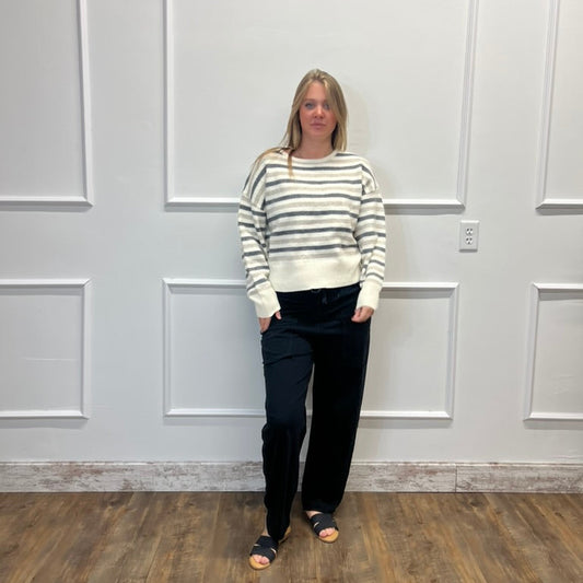 A woman stands in a room with white paneled walls and a wood floor, wearing a cream and gray striped knit sweater, black pants, and black slide sandals.