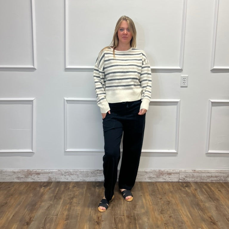 A woman stands in a room with white paneled walls and a wood floor, wearing a cream and gray striped knit sweater, black pants, and black slide sandals.