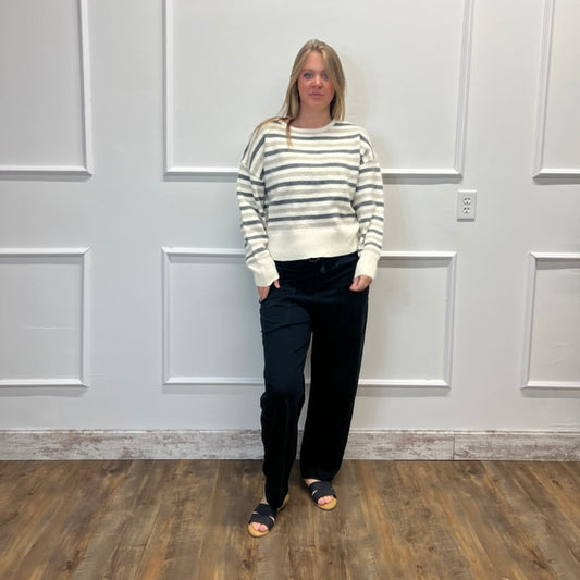A woman stands in a room with white paneled walls and a wood floor, wearing a cream and gray striped knit sweater, black pants, and black slide sandals.