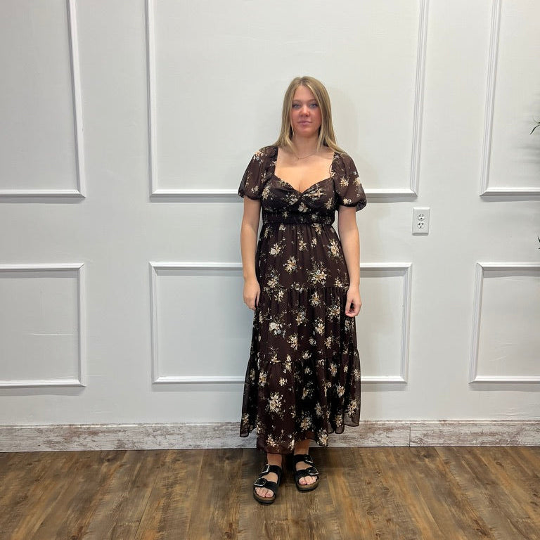 A woman stands in front of a white paneled wall wearing a dark brown midi dress with a beige floral pattern, short puff sleeves, and a sweetheart neckline. She is wearing black sandals.