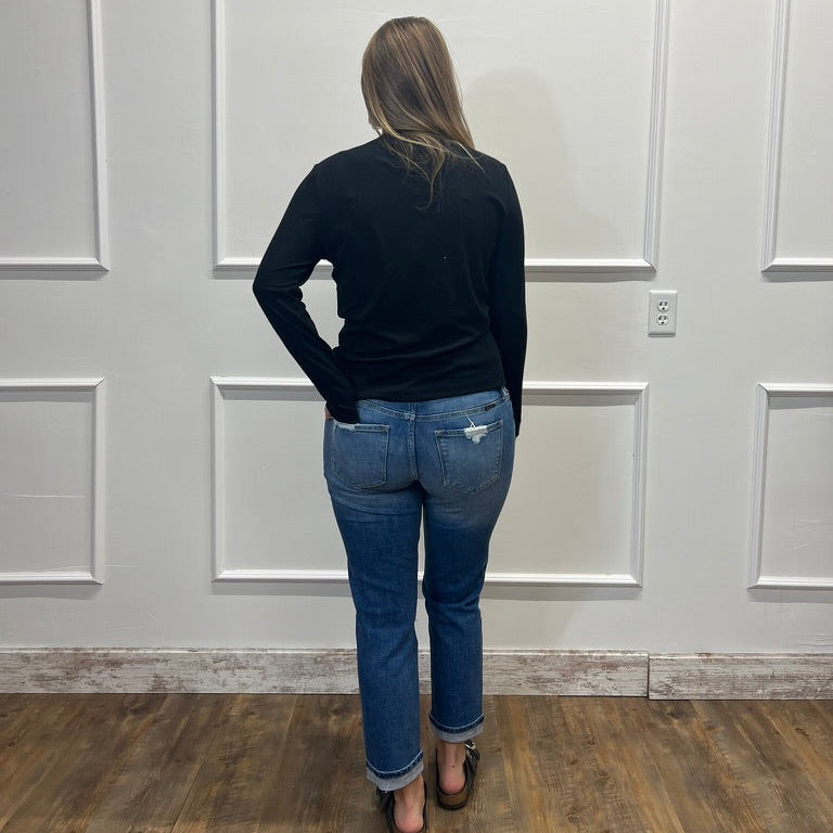 A woman viewed from behind wearing a black long-sleeve top and blue jeans, standing on a wood floor against a white paneled wall.