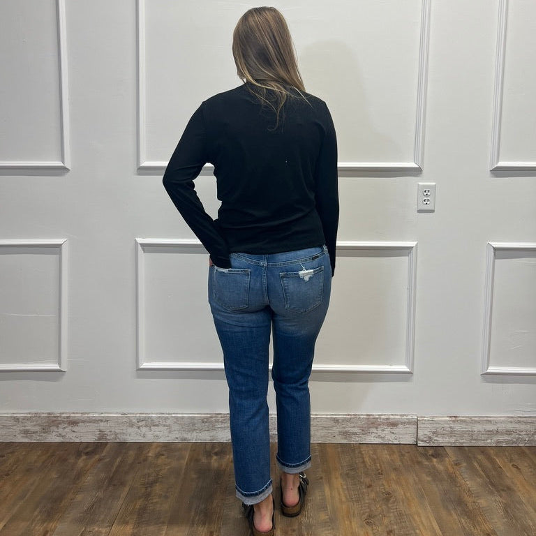 A woman viewed from behind wearing a black long-sleeve top and blue jeans, standing on a wood floor against a white paneled wall.