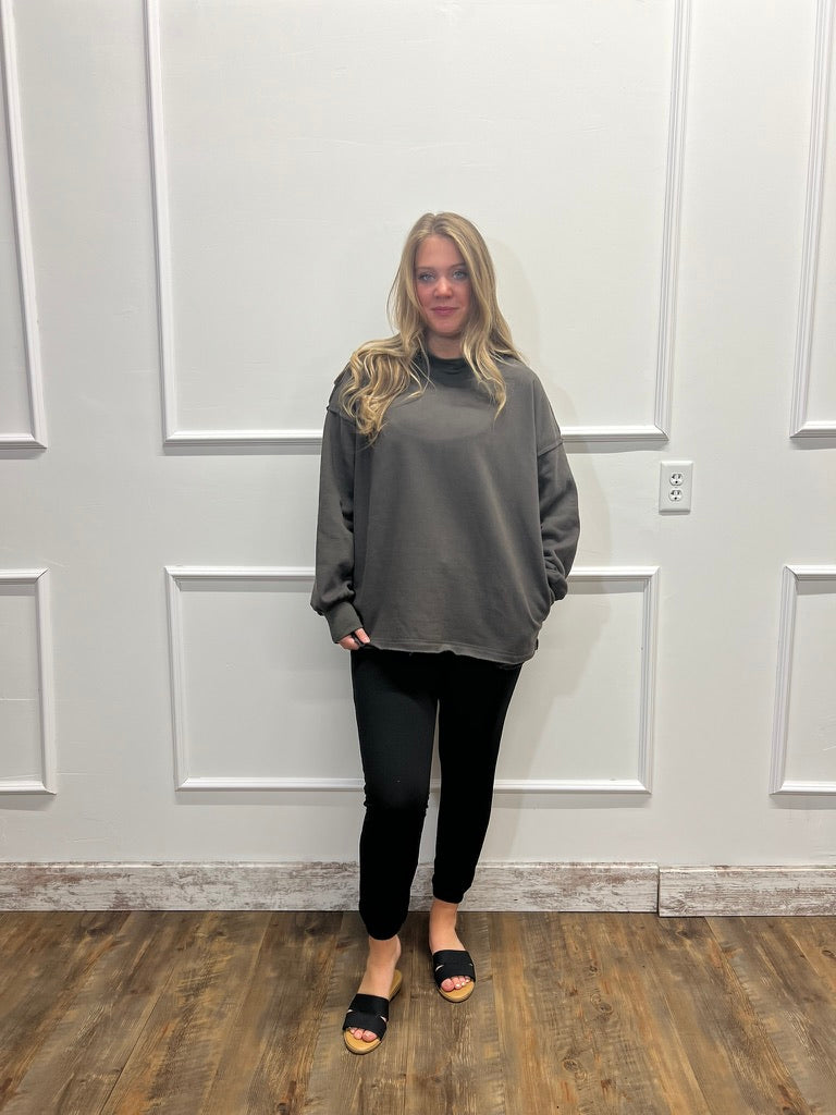 A woman stands in a room with white paneled walls and a wood floor, wearing a dark gray oversized long-sleeve top, black pants, and black slide sandals.