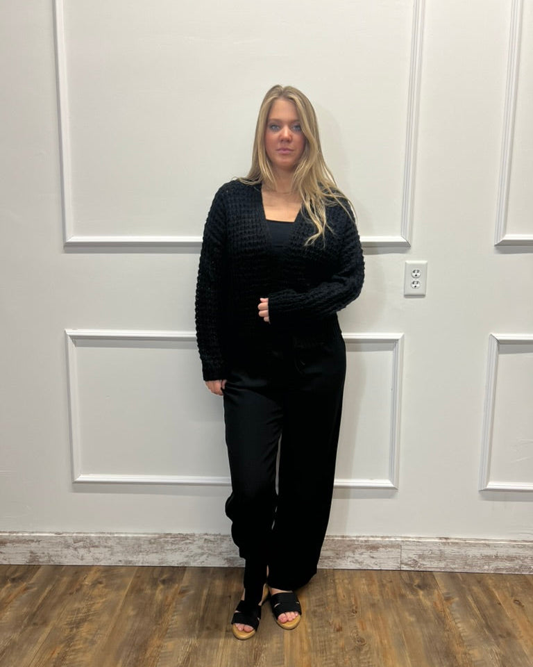A woman stands indoors against a white paneled wall wearing a black textured long-sleeve top and black wide-leg pants, with black sandals.