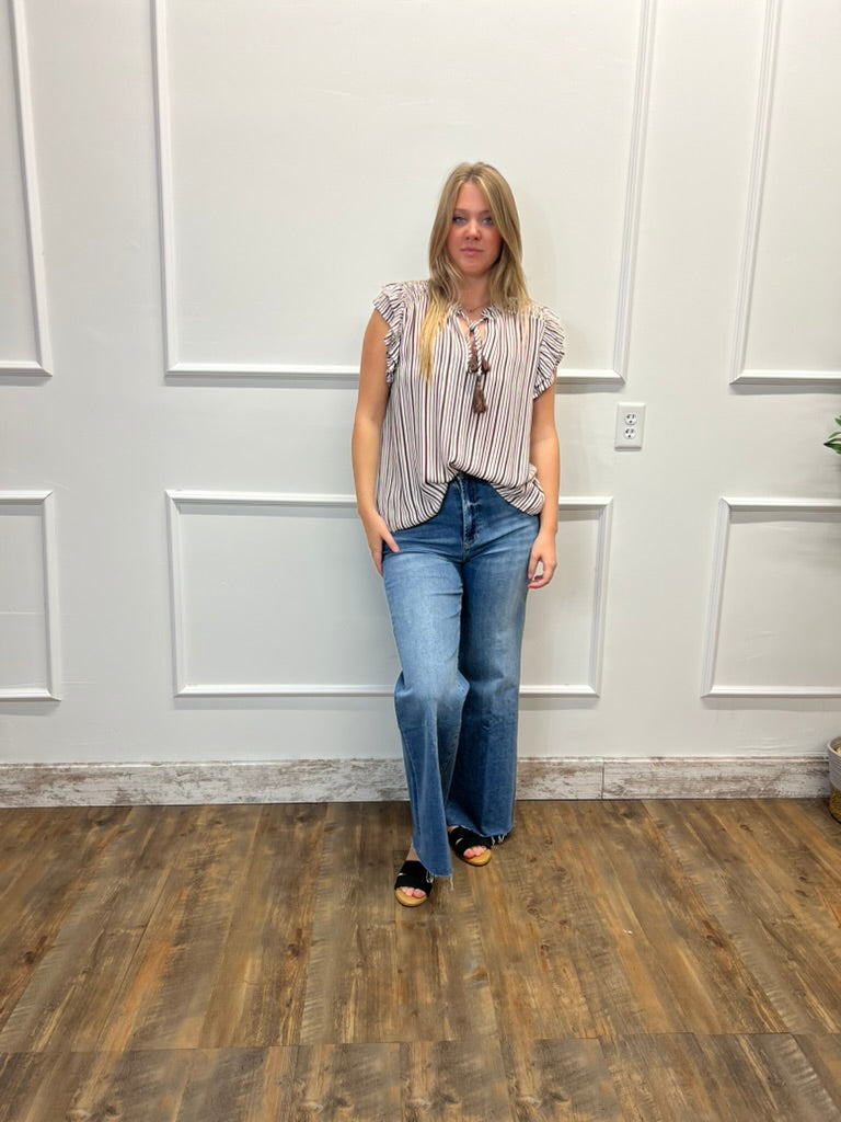 A woman stands in a room with white paneled walls and a wood floor, wearing a sleeveless striped top, blue flare jeans, and black slide sandals.