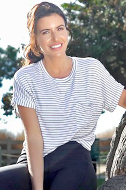 A woman smiling while wearing a white short-sleeve crewneck t-shirt with thin blue horizontal stripes and a small chest pocket, paired with dark pants, sitting outdoors next to a tree trunk.