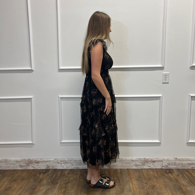 A woman stands in profile wearing a sleeveless black midi dress with a subtle floral pattern and black slide sandals, standing on a wood floor against a white paneled wall.