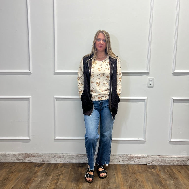 A woman stands against a white paneled wall wearing a cream floral long-sleeve top, a black zippered vest, blue jeans, and black sandals.