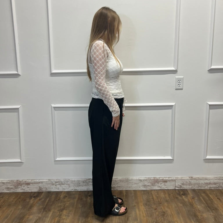 A woman stands in profile wearing a white long-sleeve mesh top and black wide-leg pants against a white paneled wall with wood flooring.