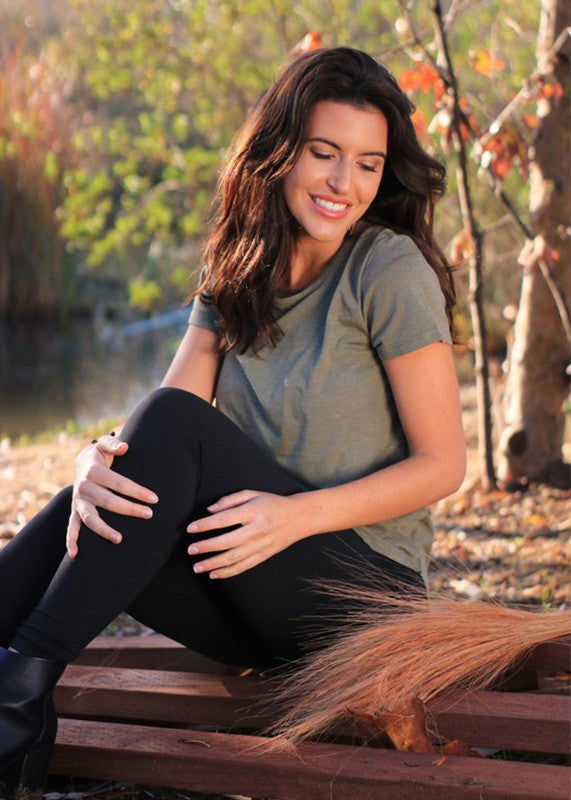A woman sitting on a wooden bench outdoors, wearing an olive green short-sleeve t-shirt and black leggings. A brown squirrel is visible in the foreground among autumn leaves.