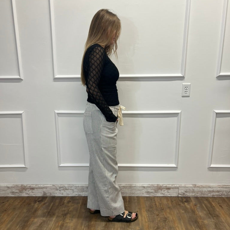 A woman stands in profile wearing a black long-sleeve fishnet top, light gray wide-leg pants, and black slide sandals, standing in front of a white paneled wall with a wood-look floor.