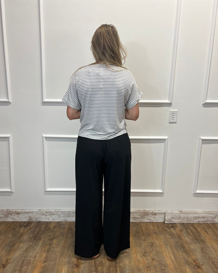 A woman viewed from behind wearing a short-sleeve gray and white striped t-shirt and black wide-leg pants, standing on a wood floor against a white paneled wall.