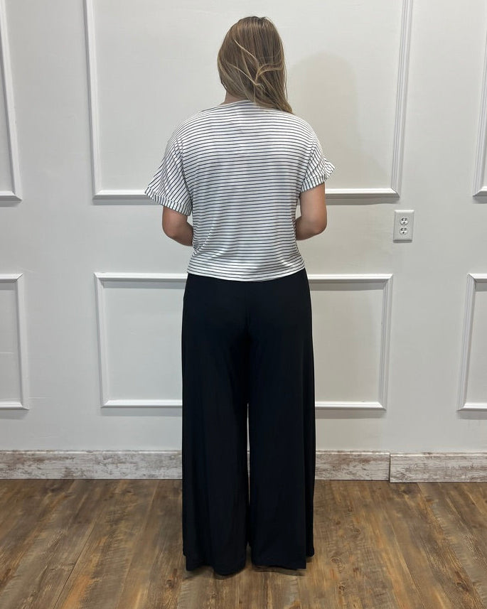 A woman stands with her back to the camera wearing a white t-shirt with thin black horizontal stripes and black wide-leg pants, standing in front of a white paneled wall on a wood floor.