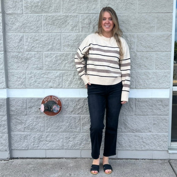 A woman stands outdoors against a light gray stone wall, wearing a cream and dark brown horizontally striped long-sleeve knit sweater, dark cropped pants, and black slide sandals.