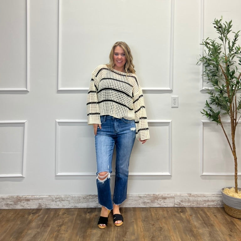 Woman wearing a striped sweater and jeans standing in a room with a white paneled wall and a plant.