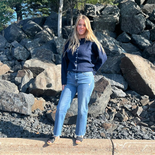 A woman stands outdoors in front of a large pile of dark rocks and trees, wearing a navy cardigan over a dark top, light blue jeans, and sandals.