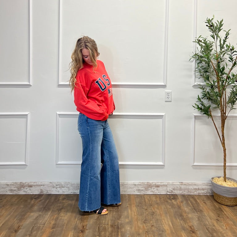 Person wearing a red sweatshirt and blue jeans standing against a white paneled wall with a plant to the right.