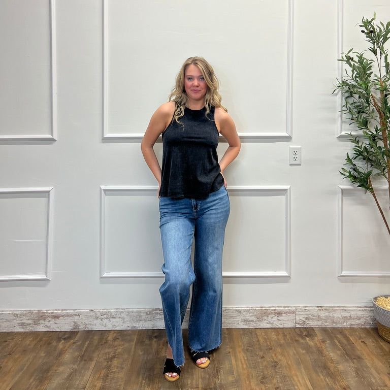 Woman wearing a black top and blue jeans standing against a white paneled wall.