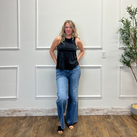 Woman wearing a black top and blue jeans standing against a white paneled wall.