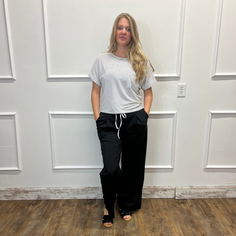 A woman stands in a room with white paneled walls and a wood floor, wearing a white short-sleeve t-shirt with thin gray horizontal stripes, black wide-leg drawstring pants, and black sandals.